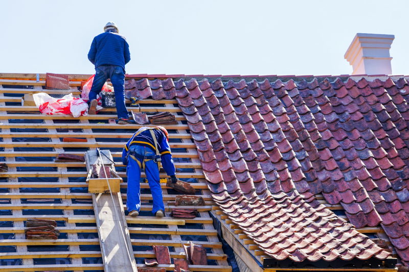 roof repair of a historic house and replacement of clay tiles Two workers in safety gear install red roof tiles on a sloped rooftop in Colorado Springs, restoring the home's exterior.