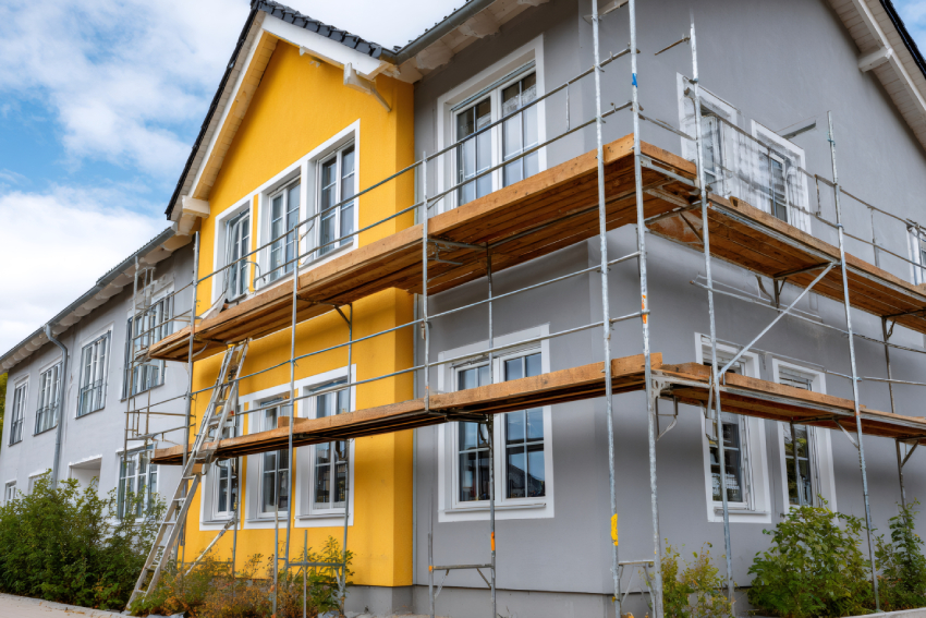 Empty unfurnished loft mansard room interior with wooden columns and wet concrete floor on roof level in black and whote style color A two-story house with gray and yellow walls under renovation in Colorado Springs, surrounded by scaffolding for exterior restoration.